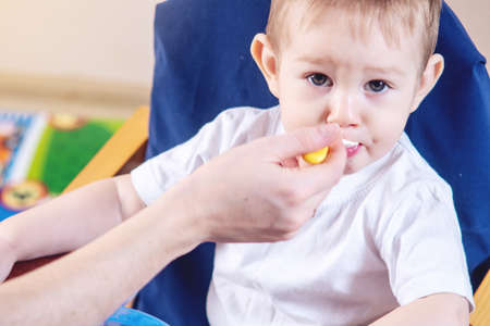 Little cute baby boy eating on a chair in the kitchen. Mom feeds holding in hand a spoon of porridgeの写真素材