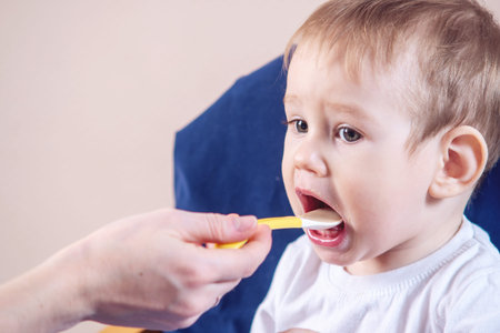 Little cute baby boy eating on a chair in the kitchen. Mom feeds holding in hand a spoon of porridgeの写真素材