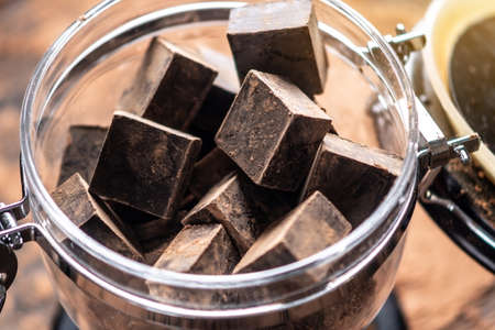 Pieces of dark bitter chocolate with cocoa powder in a glass jar on wooden background. The concept of confectionery ingredientsの写真素材