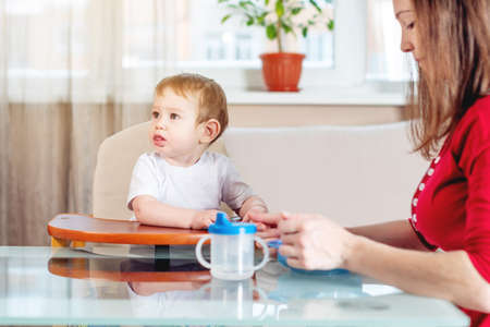Mom feeding the baby holding out her hand with a spoon of food in the kitchen. Healthy baby food. The emotions of a child while eatingの写真素材