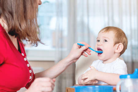 Mother feeding the baby holding out her hand with a spoon of porridge in the kitchen. Emotions of a child while eating healthy food.の写真素材