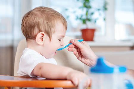 Mom feeding the baby holding out her hand with a spoon of food in the kitchen. Healthy baby food. The emotions of a child while eatingの写真素材