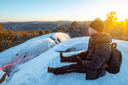 Man hiker with a backpack sitting on top of a cliff in the forests of Siberia at sunset. Beautiful snow panorama of wildlife. Freedom and winter trekkingの写真素材