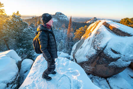 Man hiker with a backpack stands on top of a cliff in the forests of Siberia at sunset. Beautiful snow panorama of wildlife. Freedom and winter trekkingの写真素材