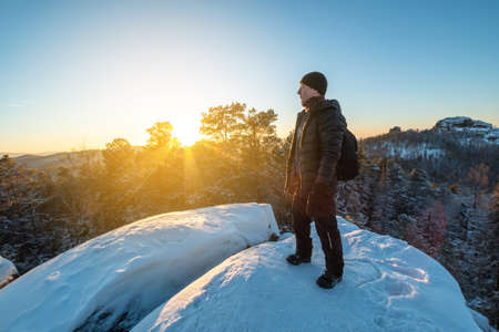 Man hiker with a backpack stands on top of a cliff in the forests of Siberia at sunset. Beautiful snow panorama of wildlife. Freedom and winter trekkingの写真素材
