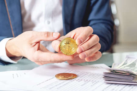 Businessman banker sitting at the table with papers holding a coin bitcoin. The concept of exchange and sale of cryptocurrency for dollarsの写真素材