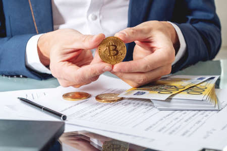 Businessman banker sitting at the table with papers holding a coin bitcoin. The concept of exchange and sale of cryptocurrency for Euroの写真素材