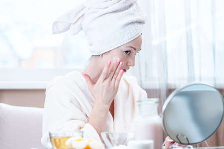 Beautiful happy young woman with a towel on her head applying cream of natural cosmetics on face moisturizing the skin. The concept of hygiene and care for the skin at homeの写真素材