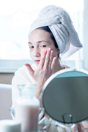 Beautiful happy young woman with a towel on her head applying cream of natural cosmetics on face moisturizing the skin. The concept of hygiene and care for the skin at homeの写真素材