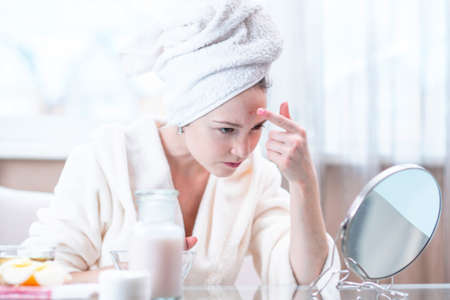 Beautiful unhappy young woman with a towel on her head detects acne on her face. Concept of hygiene and care for the skinの写真素材