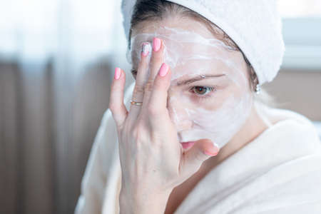 Beautiful happy young woman with a towel on her head applying cream of natural cosmetics on face moisturizing the skin. The concept of hygiene and care for the skin at homeの写真素材