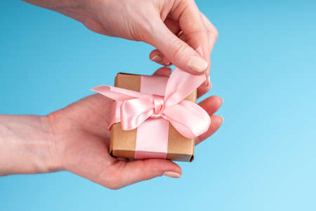 Women's hands holding gift box Packed in Kraft paper with pink ribbon on blue background. Concept holiday card for Valentine's day and women's dayの写真素材