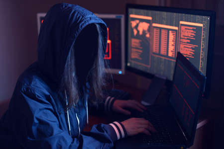 Anonymous hacker girl with no face typing the code tries to hack the system and steal accesses on the background screens in neon light. The concept of cybersecurityの写真素材