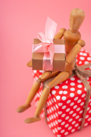 Wooden toy model holding gifts sitting on a red polka dot box on pink background. Concept holiday card for Valentine's day and women's dayの写真素材