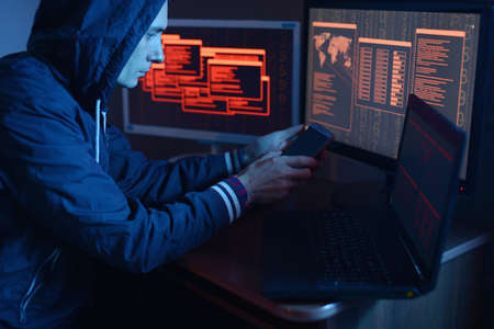 A male hacker in a hood typing program code while committing a cybercrime hacking a system firewall on the background of screens in the dark under neon light. Cyber security conceptの写真素材
