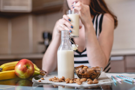 Woman drinking organic almond milk holding a glass in her hand in the kitchen. Bottle and fruit on the table. Diet healthy vegetarian productの写真素材