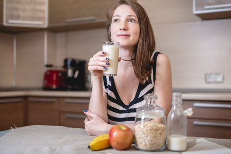 Happy young woman holding a glass of organic oat milk in the kitchen. Bottle and fruit on the table....