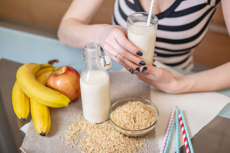 Girl drinking organic rice milk holding a glass Cup in her hands in the kitchen. Bottle and fruit on the table. The view from the top. Diet healthy vegetarian productの写真素材