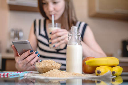 Happy young drinking organic rice milk while holding the phone in the kitchen. Bottle and fruit on the table. Diet healthy vegetarian productの写真素材