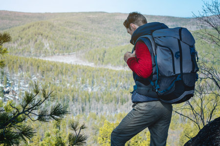 Man tourist hiker with a large backpack stands on a rock cliff and looks at the sprawling green...