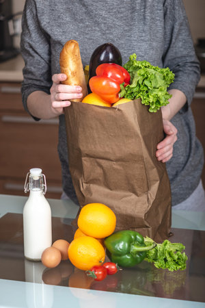 A woman unpacks a full paper bag with products on the background of the kitchen. Healthy and fresh...