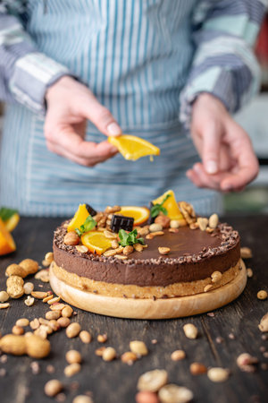 Confectioner decorates chocolate cake with orange and mint leaves with peanuts and nuts. Healthy raw...