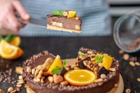 Pastry chef in hand holding a piece of chocolate cake with orange, mint leaves, peanuts and nuts. Healthy raw gluten-free and milk-and flour-free desserts for vegan foodの写真素材