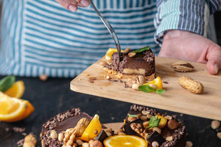 Hand breaks with a fork a piece of chocolate cake with orange, mint leaves, peanuts and nuts. Concept healthy raw gluten-free and milk-and flour-free desserts for vegan foodの写真素材