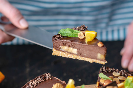 Pastry chef in hand holding a piece of chocolate cake with orange, mint leaves, peanuts and nuts. Healthy raw gluten-free and milk-and flour-free desserts for vegan foodの写真素材