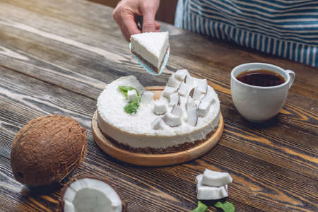 Confectioner holding a piece of coconut raw cake with white pulp and mint on a wooden background. Healthy fresh summer vegan dessert. Gluten and sugar free foodの写真素材