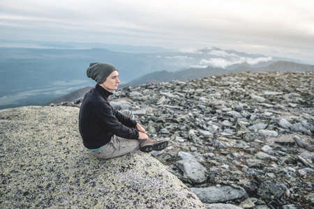 Man hiker sitting in a yoga pose at the peak of the mountain in the summer. Meditation after a long climb on a mountain background of cloudy sky in the eveningの写真素材