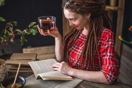 Beautiful woman reading a romantic book in a cozy warm atmosphere enjoying the autumn mood. Rustic table with yellow flowers and a stack of booksの写真素材