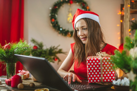 Beautiful woman in a red dress is holding a gift box and ordering online purchases on her laptop. Online shopping for Christmas holidays.の写真素材