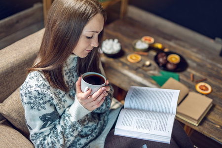 Young woman in a sweater reading a book with a mug of coffee in the evening in a warm Christmas...