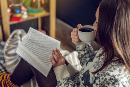 Young woman in a sweater reading a book with a mug of coffee in the evening in a warm Christmas atmosphere. Cozy new year moodの写真素材