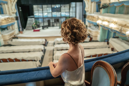 A beautiful woman in a dress alone on the balcony of a classical empty theater looks at the stage...