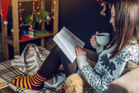 Young girl in a sweater reading a book with a mug of coffee in the evening in a warm Christmas atmosphere. Cozy new year moodの写真素材