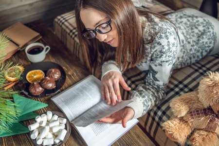 Young girl in a sweater reading a book with a cup of coffee in the evening in a warm Christmas...