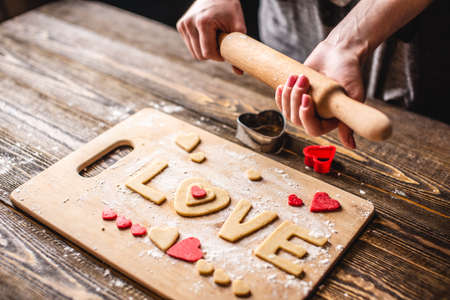 Cooking cookies from the dough in the shape of a heart and the word love on dark wooden background. Baking for Valentine's day and a romantic dateの写真素材