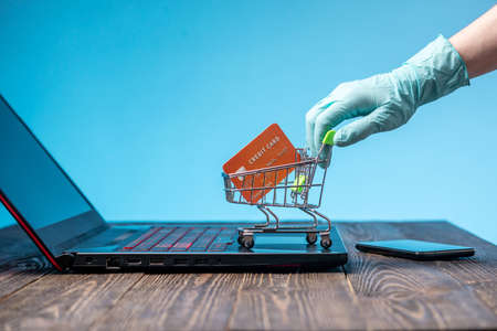 A hand in a sterile medical glove holds a shopping cart with a credit card for online purchases. Concept of internet purchasing important life support products during the coronavirus pandemicの写真素材