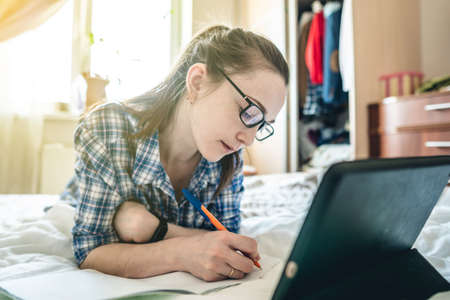A young woman student is passing online lesson on the Internet at home lying on a bed. Remote learning in schools and universities during the Covid-19 pandemicの写真素材