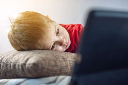 A preschool toddler child watches cartoons on a tablet on the internet while lying on a pillow on the floor. Gadgets and modern entertainment for children.の写真素材