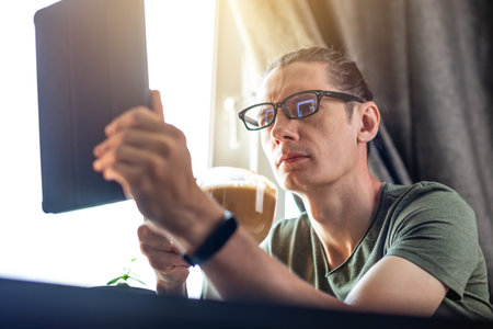 A young man in the surfing in the Internet and watching videos using a tablet at the window in flat....