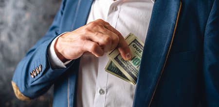 Man in a suit putting a wad of hundred-dollar bills in the inside pocket of his jacket. Close up. The concept of corruption and bribery officialsの写真素材