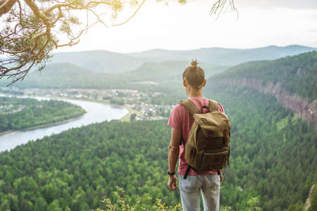 A tourist traveler with a backpack is standing on the edge of a cliff and is looking on a green valley with the river. The concept of freedom and unity with natureの写真素材