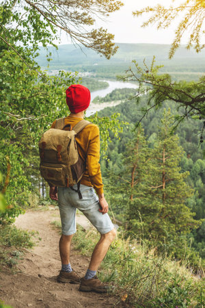 A tourist traveler with a backpack and a red hat is standing on the edge of a cliff and is looking...