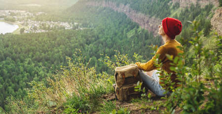 A tourist traveler with a backpack and a red hat is sitting on the edge of a cliff and is looking on a green forest valley with the river. The concept of freedom and unity with natureの写真素材