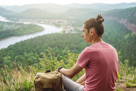 A tourist traveler with a backpack is sitting on the edge of a cliff and is looking on a green forest valley with the river. The concept of freedom and unity with natureの写真素材