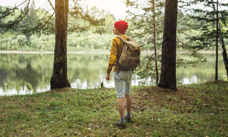 A tourist with a backpack and a red hat is walking in the forest among the trees. The concept of active trekking and unity with natureの写真素材