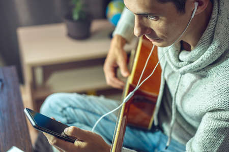 A young musician is learning to play acoustic guitar in an online lesson using a phone app. A man is studying courses at home in a room by the light of a lampの写真素材